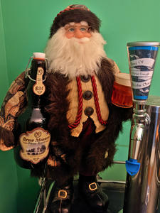 A Santa statuette on top of a kegerator, next to the chrome draft tower. Santa is a brewer, holding a disproportionately large flip top bottle filled with white string lights with a label reading Bew Master's Special Reserve.